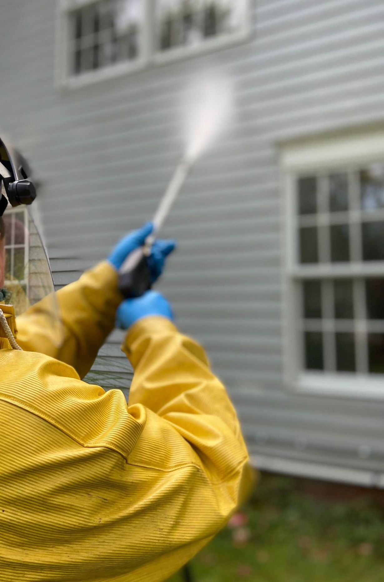 Person cleaning house siding with a pressure washer, wearing protective gear in a yellow raincoat.